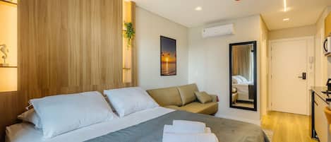 A hotel room with a bed, a sofa, a mirror, a desk, and an air conditioning unit.