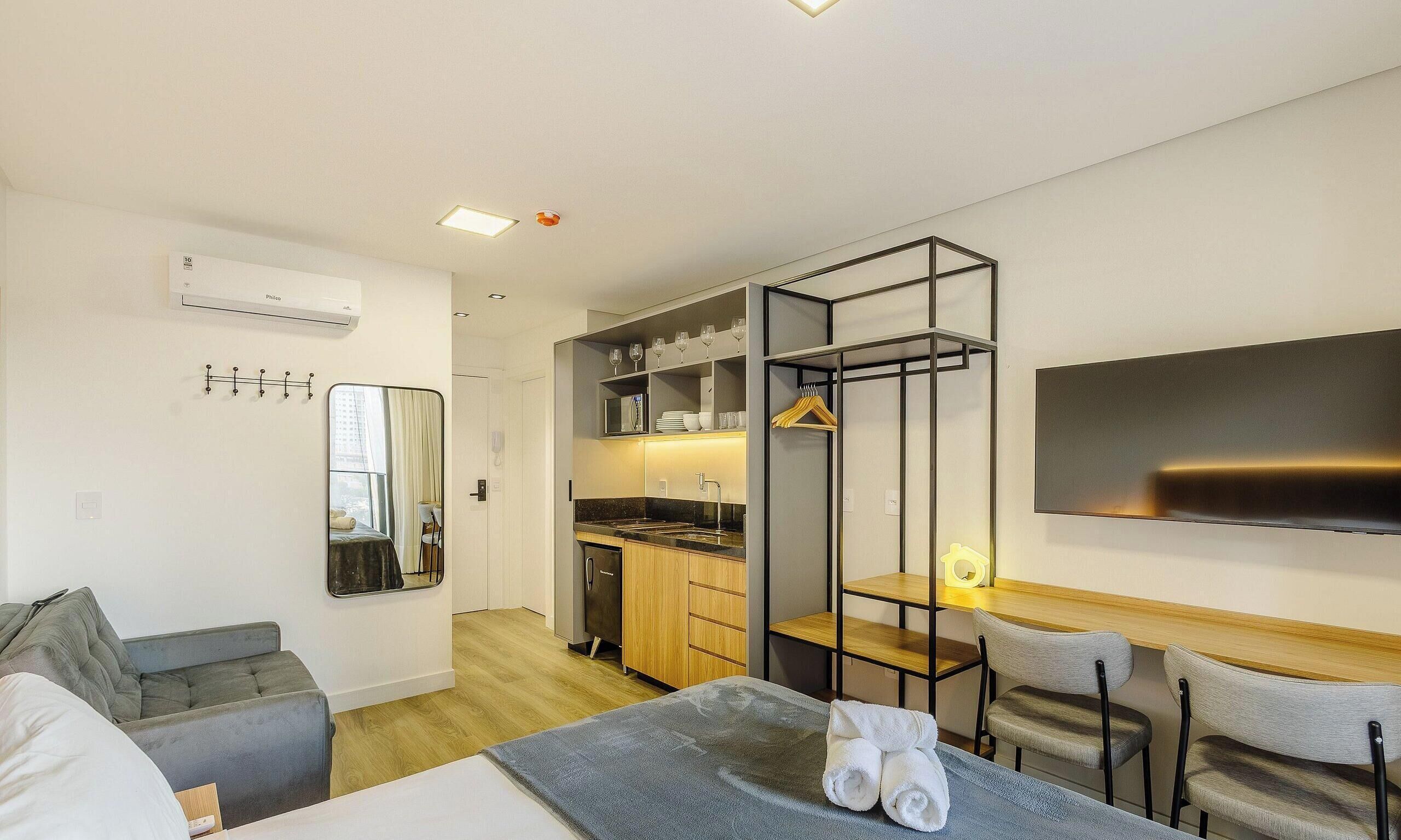 Una habitación de hotel moderna con cama, escritorio, silla, televisor, espejo, armario y kitchenette.
