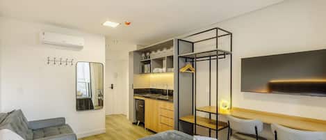 A modern hotel room with a sofa, a dining table with chairs, a kitchen area, and a flat-screen TV mounted on the wall.