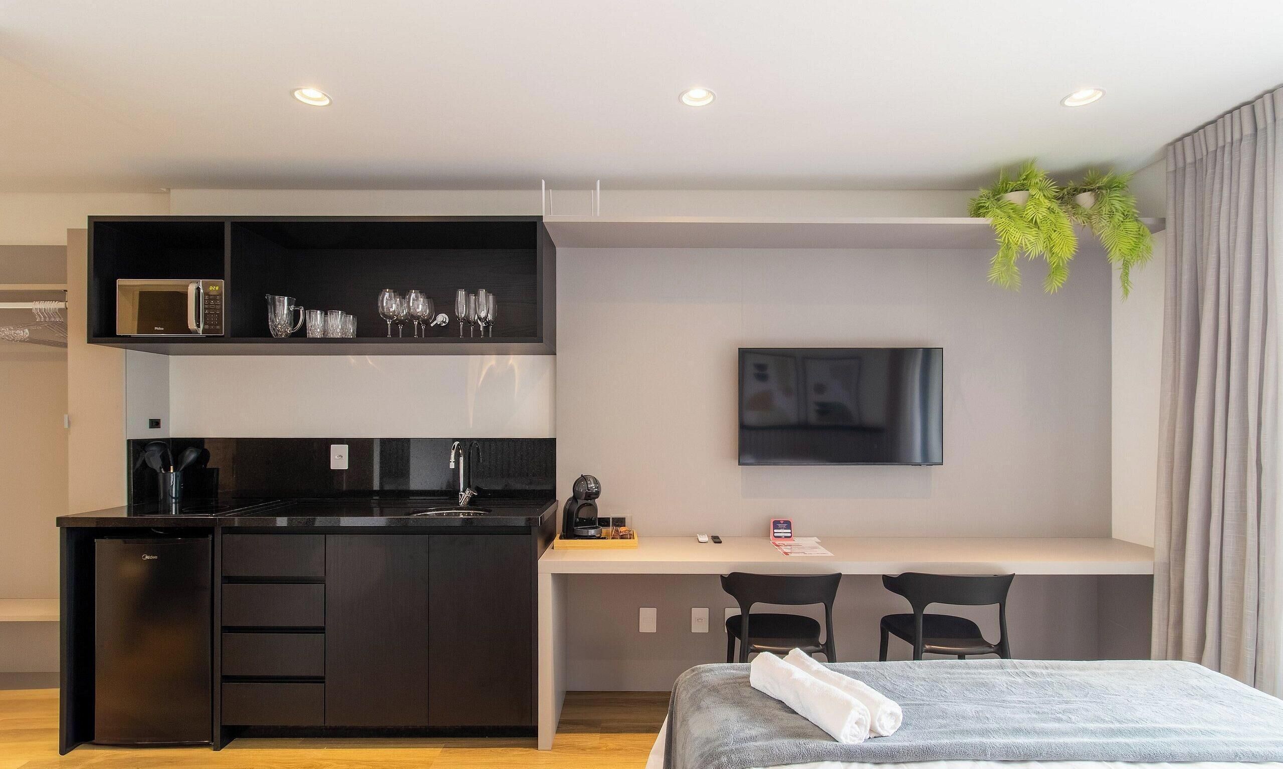 Una habitación de hotel moderna con kitchenette, un televisor de pantalla plana, una cama con manta gris y un pequeño escritorio.