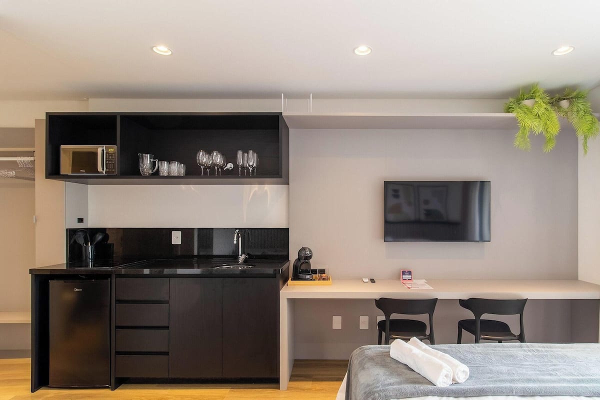 Una habitación de hotel moderna con kitchenette, un televisor de pantalla plana, una cama con manta gris y un pequeño escritorio.