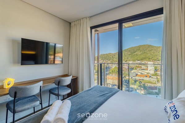 A hotel room with a bed, two chairs, a desk, a TV, and a large window with a city view. - Studios With Side Sea View at Penha Spot PSO - L (Penha)