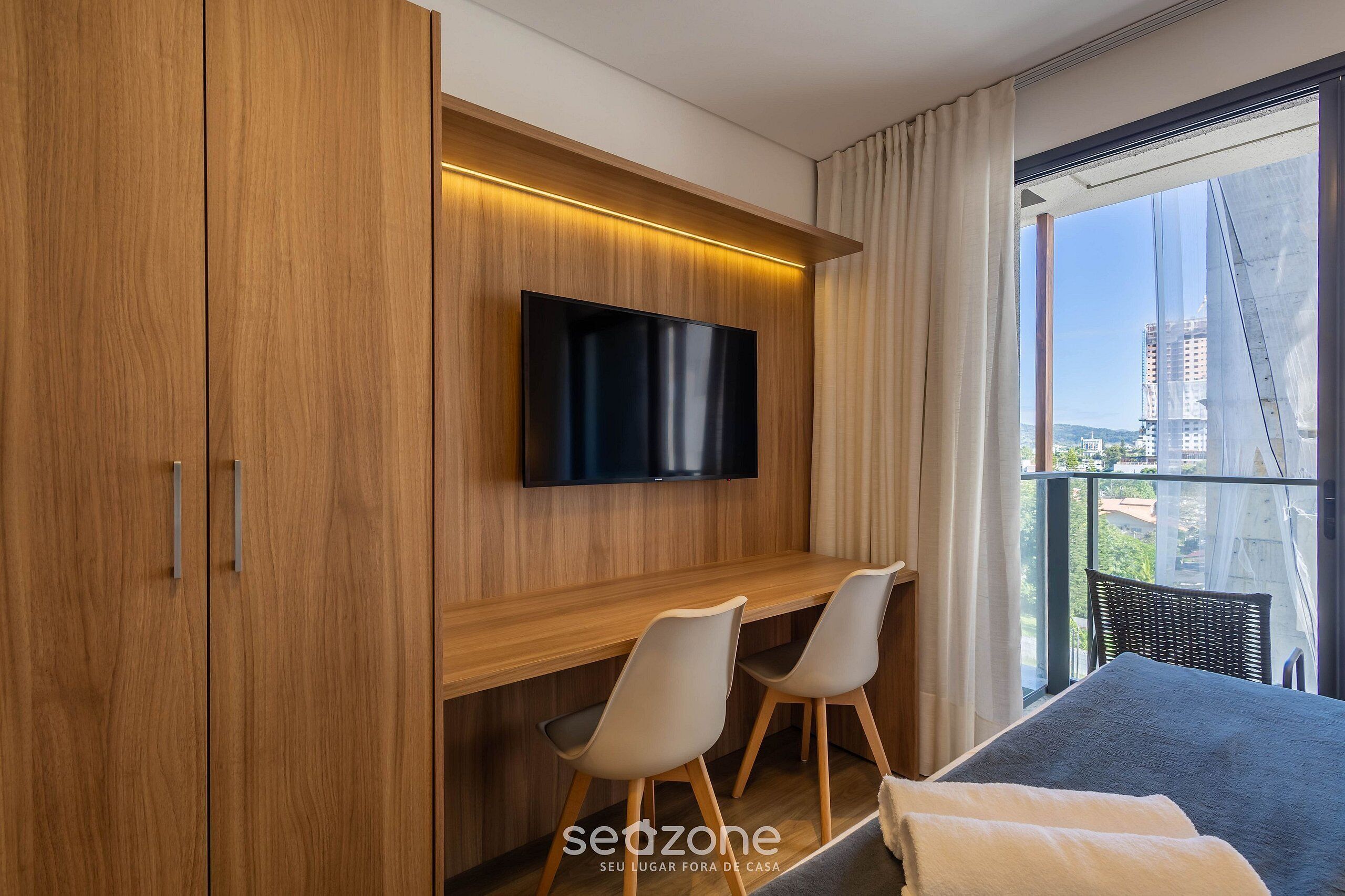 Una habitación de hotel moderna con una televisión montada en una pared de madera, un escritorio con dos sillas, una cama con un edredón oscuro y un amplio ventanal con vistas a la ciudad.