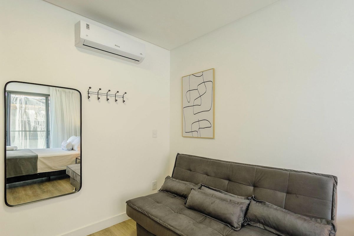 Una habitación con una cama, un sofá gris, aire acondicionado de pared, un espejo y una obra de arte abstracta enmarcada.