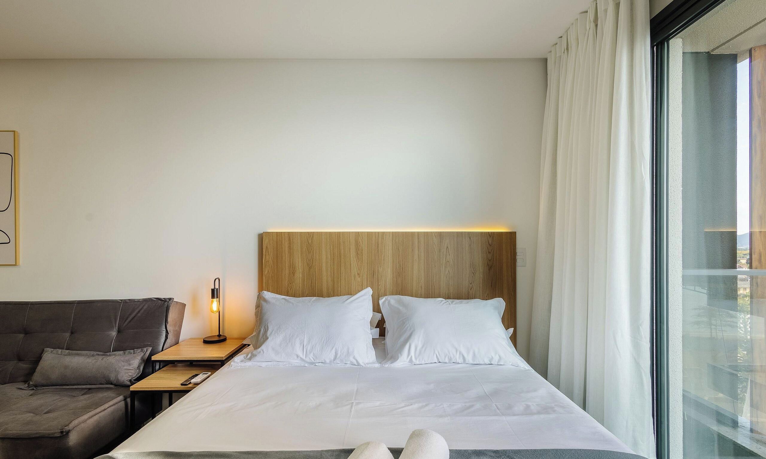Habitación de hotel con cama, mesita de noche, lámpara, sofá y un gran ventanal con cortinas.