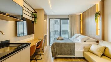 A modern hotel room with a bed, a sofa, a desk, and a kitchen area.