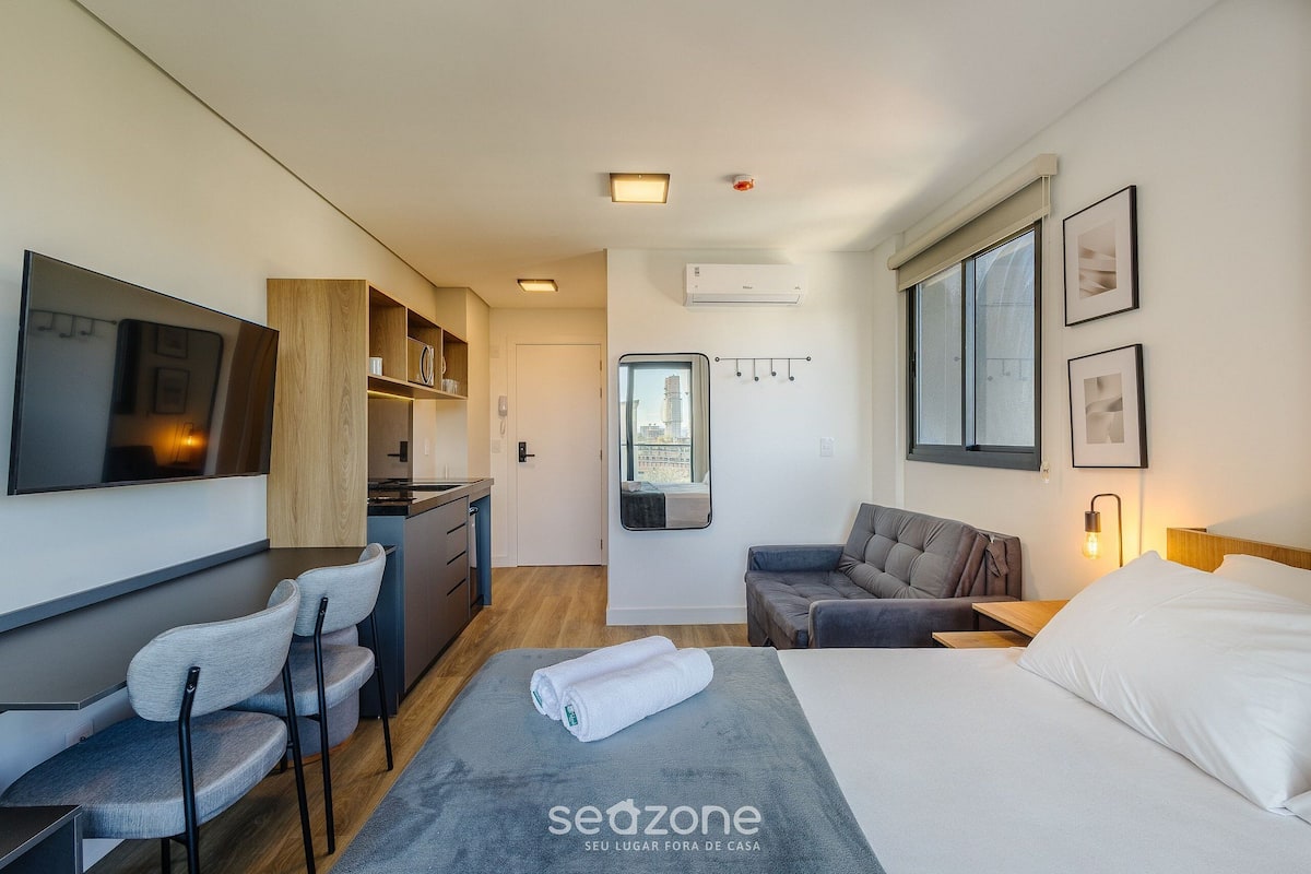 Una habitación de hotel moderna con cama, zona de estar, kitchenette y un amplio ventanal.
