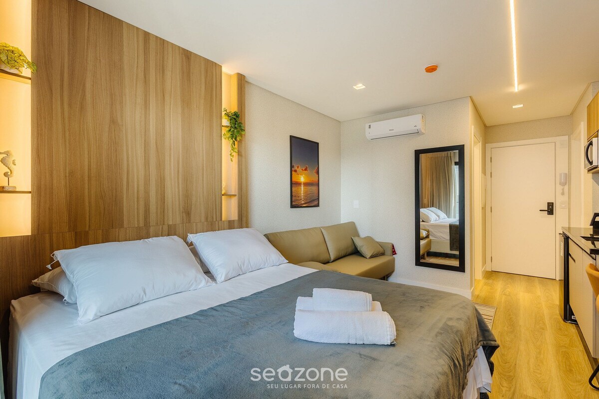 A hotel room with a bed, a sofa, a mirror, a desk, and an air conditioning unit.