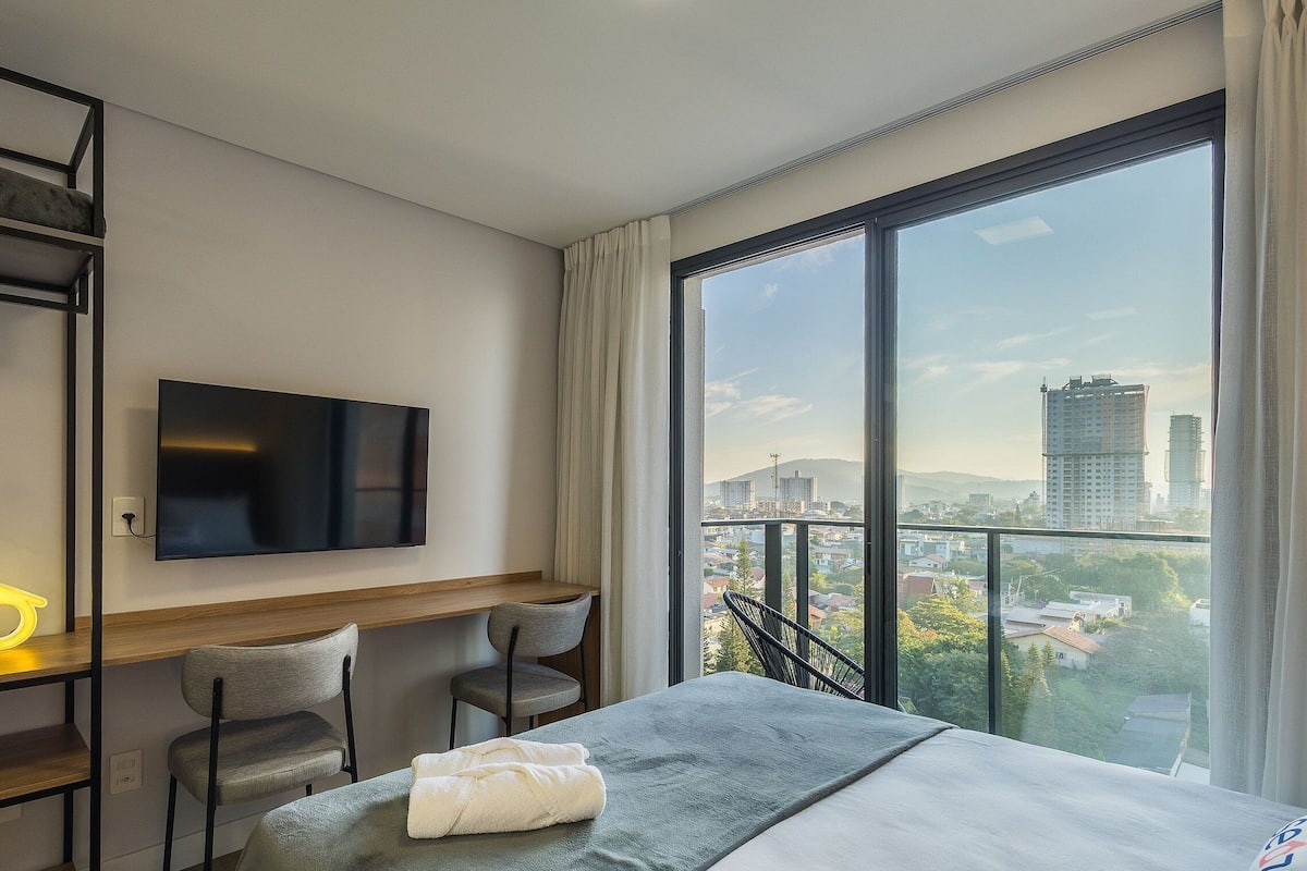 Habitación de hotel con cama, escritorio, dos sillas, televisión y un amplio ventanal con vista a la ciudad.