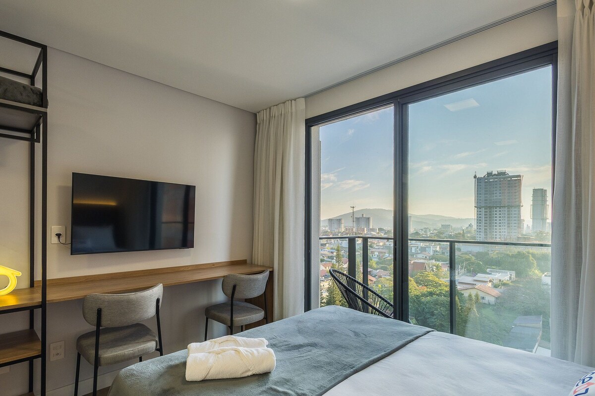 A hotel room with a bed, a desk, two chairs, a TV, and a large window with a city view.