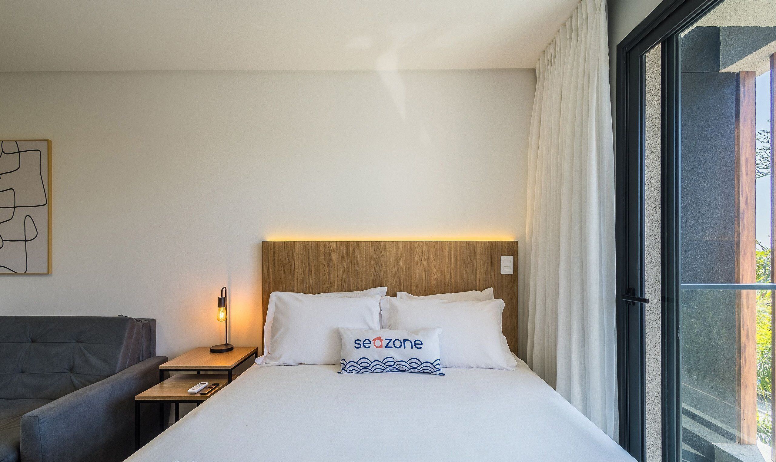 Una habitación de hotel con una cama, una mesita de noche con lámpara, un sofá y una ventana con cortinas.