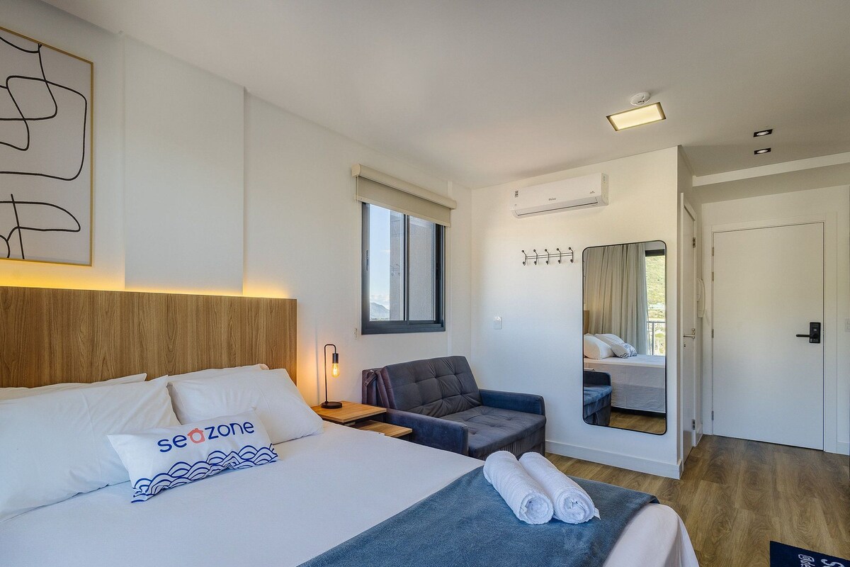 A modern hotel room with a large bed, a seating area, a window, and a mirror.