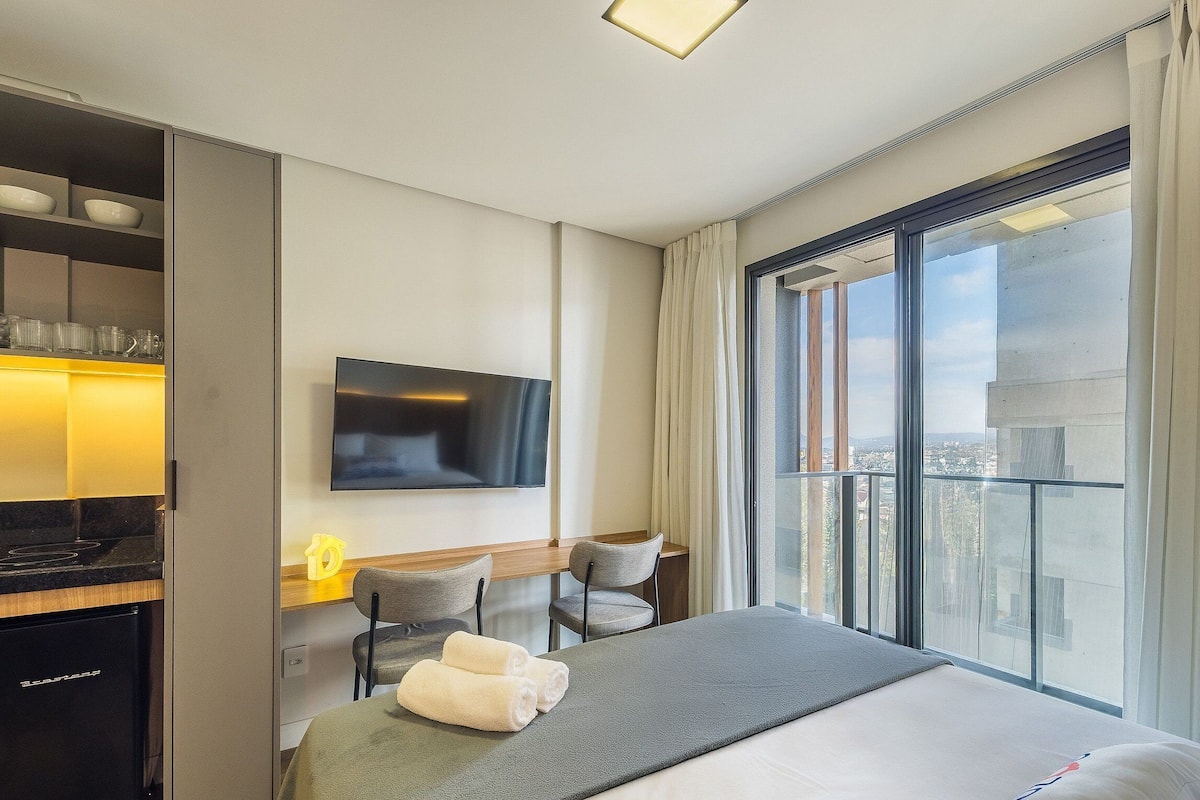 Habitación de hotel con cama, escritorio con silla, televisión de pantalla plana y ventana grande con cortinas.