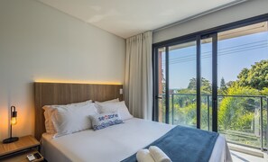 Habitación de hotel con cama, mesita de noche con lámpara, control remoto y vista a zonas verdes a través de amplios ventanales.