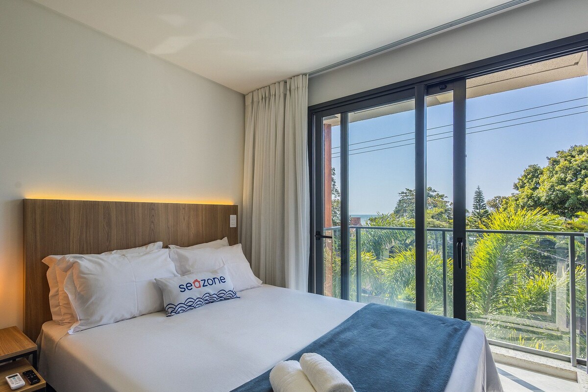 Habitación de hotel con cama, mesita de noche con lámpara, control remoto y vista a zonas verdes a través de amplios ventanales.