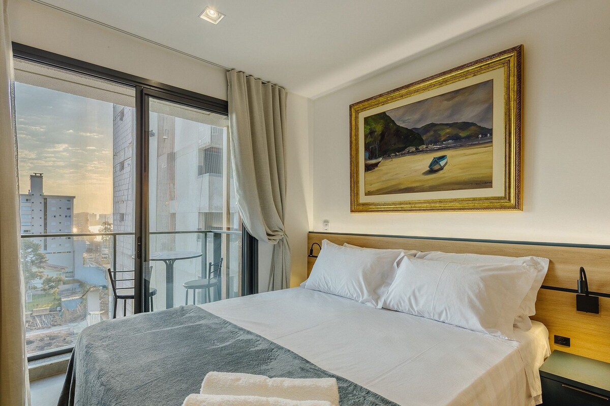 A hotel room with a large bed, a painting on the wall, and a view of buildings through the window.