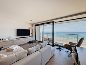 A modern living room with a large TV, comfortable seating, and a view of the beach.