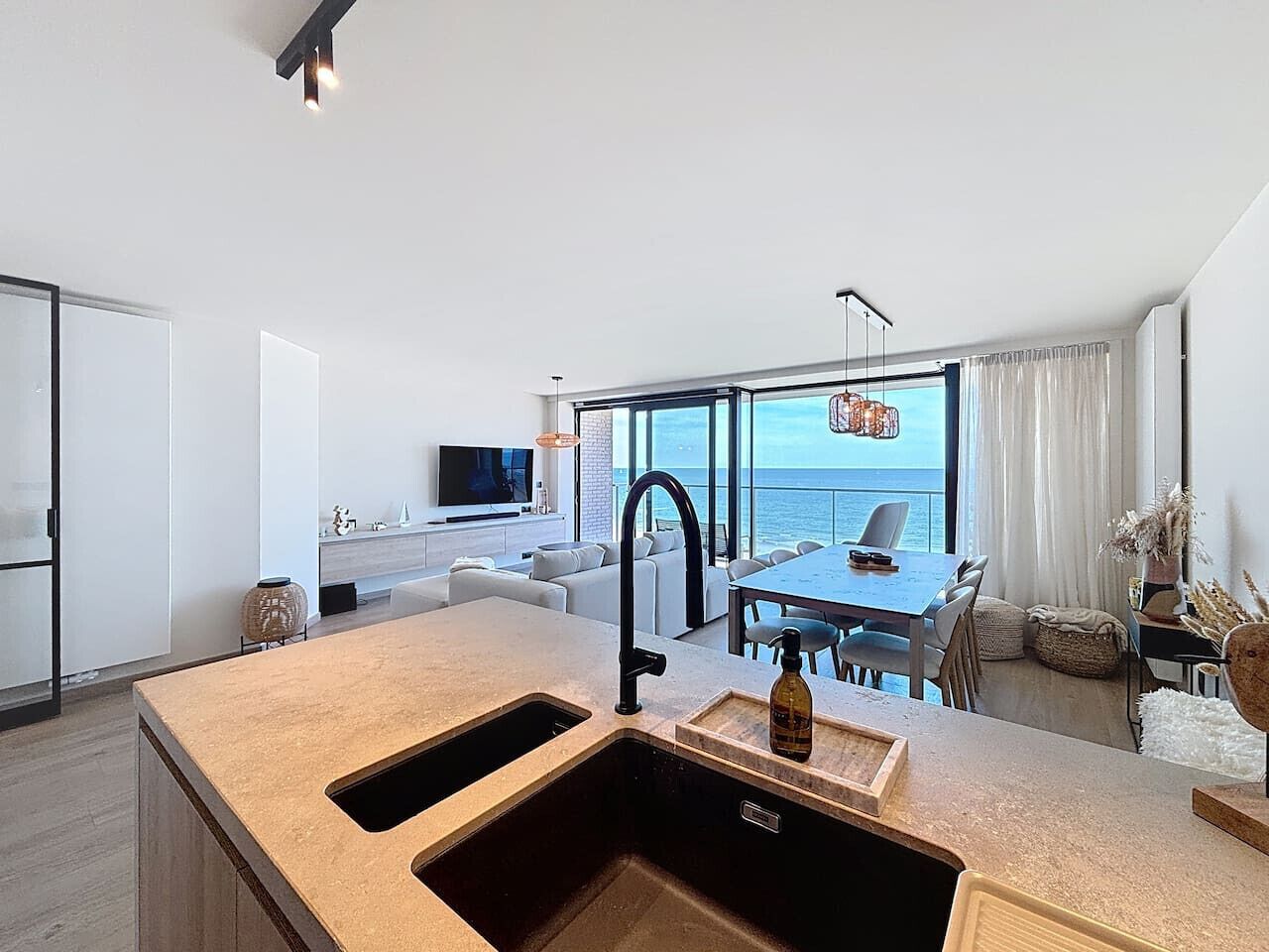 Une cuisine moderne dotée d’un grand îlot central, un coin repas offrant une vue sur la mer, et un espace de vie avec un canapé et une télévision.