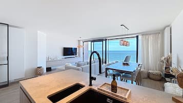 Une cuisine moderne dotée d’un grand îlot central, un coin repas offrant une vue sur la mer, et un espace de vie avec un canapé et une télévision.