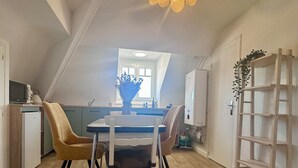 Uma sala de jantar com uma mesa, cadeiras, um vaso com flores, um lustre e uma cozinha ao fundo.