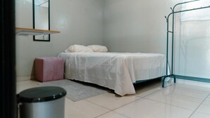 A small, clean room with a bed, a pink ottoman, a trash can, and a metal rack. - All inclusive apartment (San Salvador)