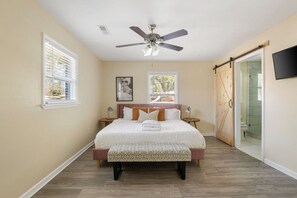 A bedroom with a bed, bedside tables, a ceiling fan, a window, and a sliding door leading to a bathroom. - Pool Home - Perfect for Families - 4 Bedrooms (Hanahan)