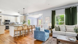 Una cocina moderna con armarios blancos, una isla central con taburetes de bar, un sillón azul y un sofá gris en una sala de estar con un ventanal y cortinas verdes.