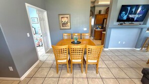 A dining area with a wooden table and chairs, a television mounted on the wall, and a kitchen in the background.
