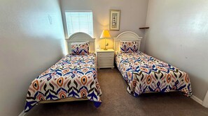 A bedroom with two single beds, a nightstand with a lamp, a window with blinds, and a framed picture on the wall.