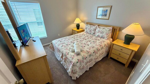 Un dormitorio con una cama, dos mesitas de noche con lámparas, un escritorio con ordenador y una ventana con persianas.