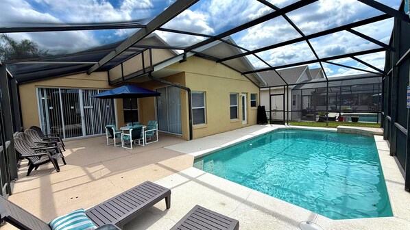 An outdoor swimming pool area with a covered patio, lounge chairs, and a table.