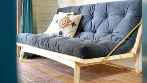 Ein Sofa mit Holzrahmen, grauem Kissen und einem gemusterten Kopfkissen, platziert in einem Raum mit Holzboden und einem fenster mit Vorhängen.