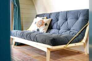 Un canapé à ossature en bois, doté de coussins gris et d’un coussin à motifs, placé dans une pièce au plancher en bois et ornée d’une fenêtre avec des rideaux.