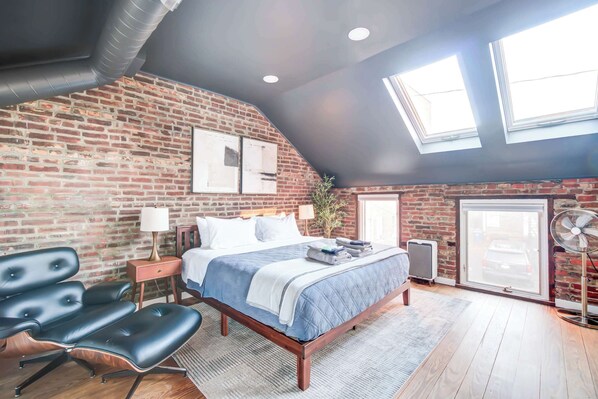 Une chambre avec un mur en briques, un lit recouvert d’une courtepointe bleue, un bureau avec une lampe et un ventilateur.