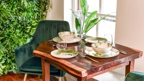 Una mesa de comedor dispuesta con platos, cuencos y cubiertos blancos, rodeada de sillas verdes y una planta en maceta.