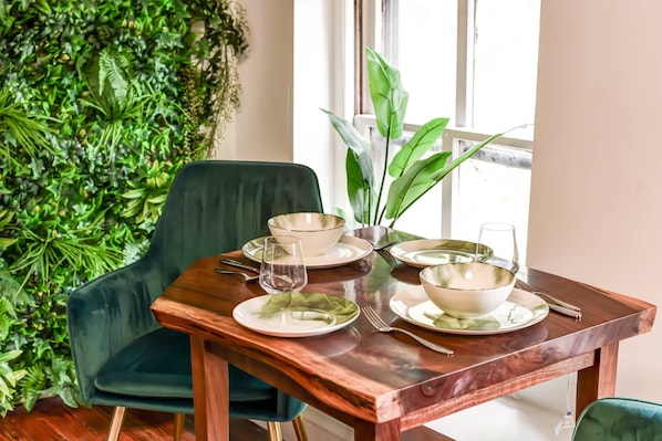 A dining table set with white plates, bowls, and cutlery, surrounded by green chairs and a potted plant.
