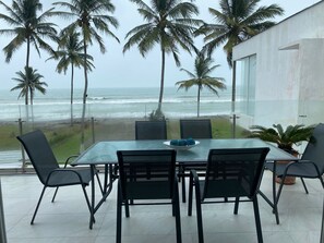 A dining area with a table and chairs, overlooking a beach with palm trees and the ocean.