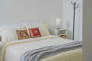 Een netjes opgemaakt bed met een witte deken en twee decoratieve kussens, een nachtkastje met een lamp, en een staande vloerlamp.