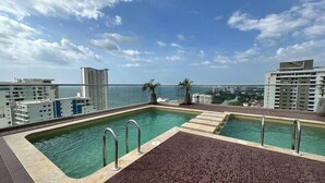 Ein Pool auf dem Dach mit klarem, blauem Wasser, umgeben von einer Holzterrasse und Metallgeländern, mit Blick auf eine Stadtlandschaft und das Meer.