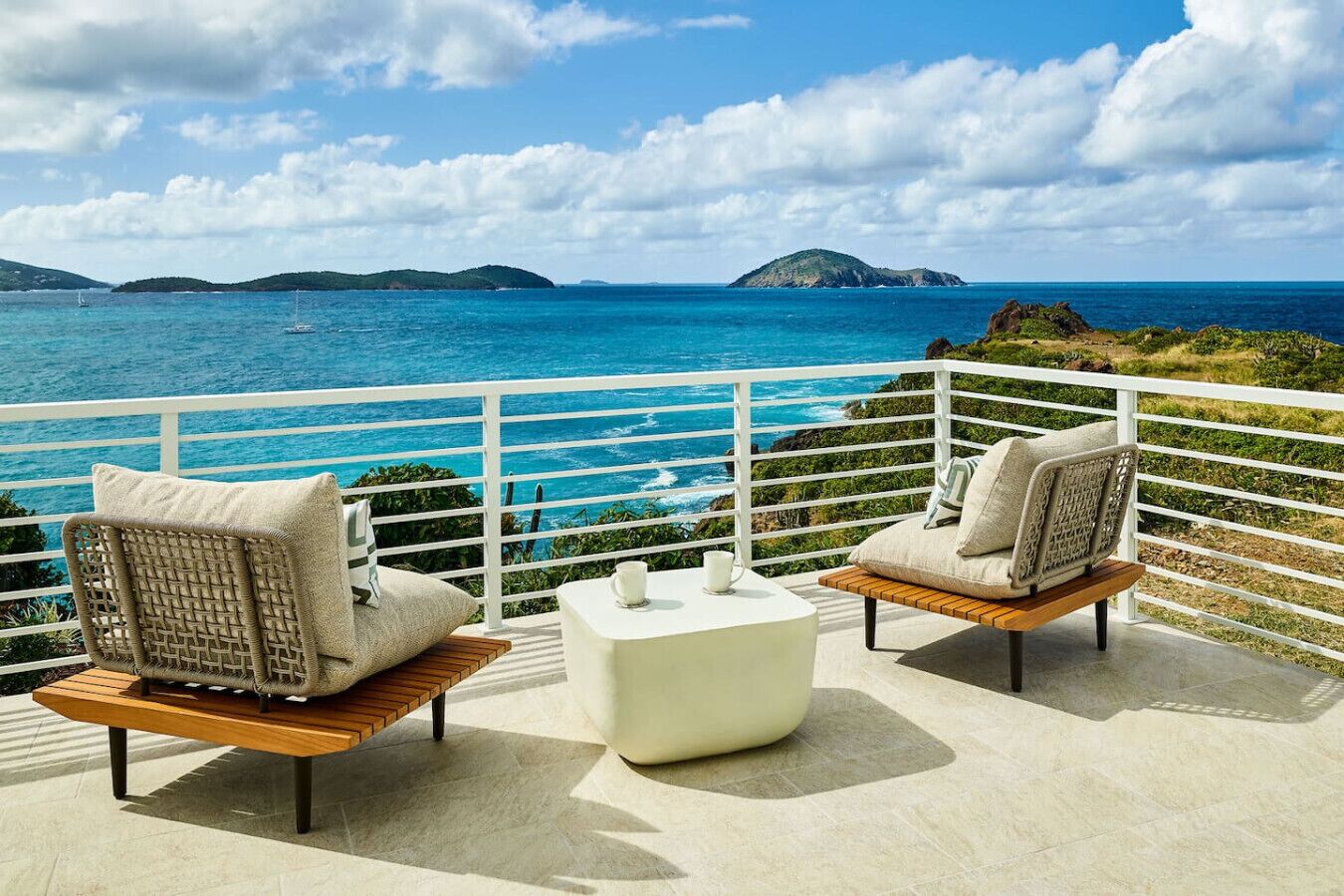 A balcony with two wicker chairs and a small white table, overlooking a turquoise sea and distant islands.