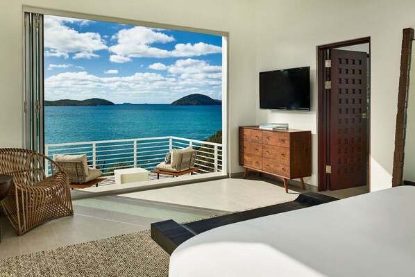 Un dormitorio con un ventanal grande que ofrece vistas panorámicas del océano, una cama, un aparador y un televisor en la pared.
