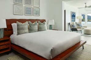 Ruime slaapkamer met een groot bed, houten hoofdbord en meerdere kussens. Er zijn nachtkastjes met lampen, een uitzicht op de omgeving door grote ramen, en een zithoek met stoelen en een tafel.