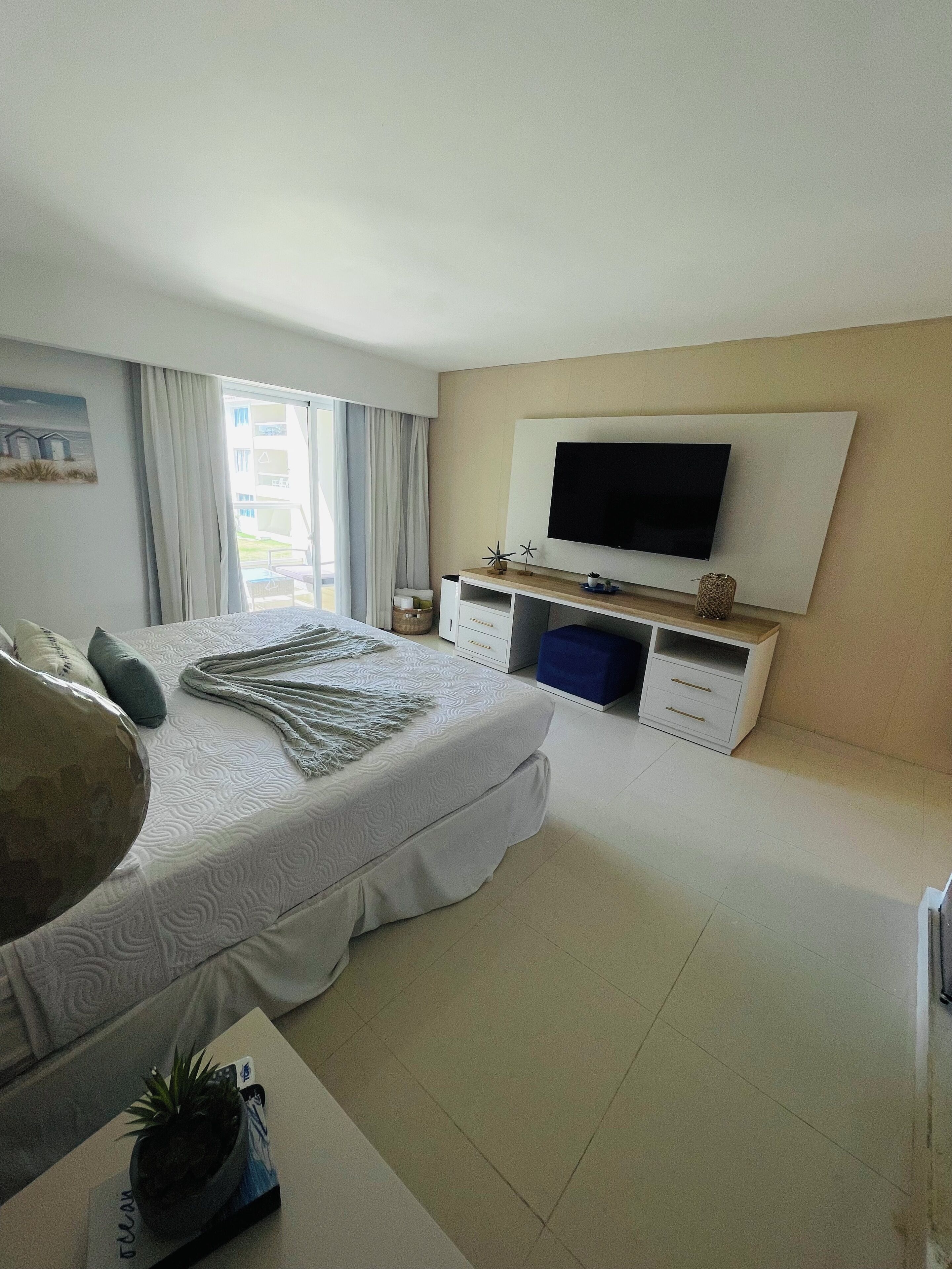 A modern hotel bedroom with a large bed, a flat-screen TV mounted on the wall, and a balcony with curtains.