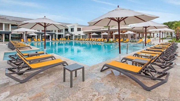 Een zwembadterras met ligbedden en parasols, een heldere lucht, en een gebouw met een bord waarop 