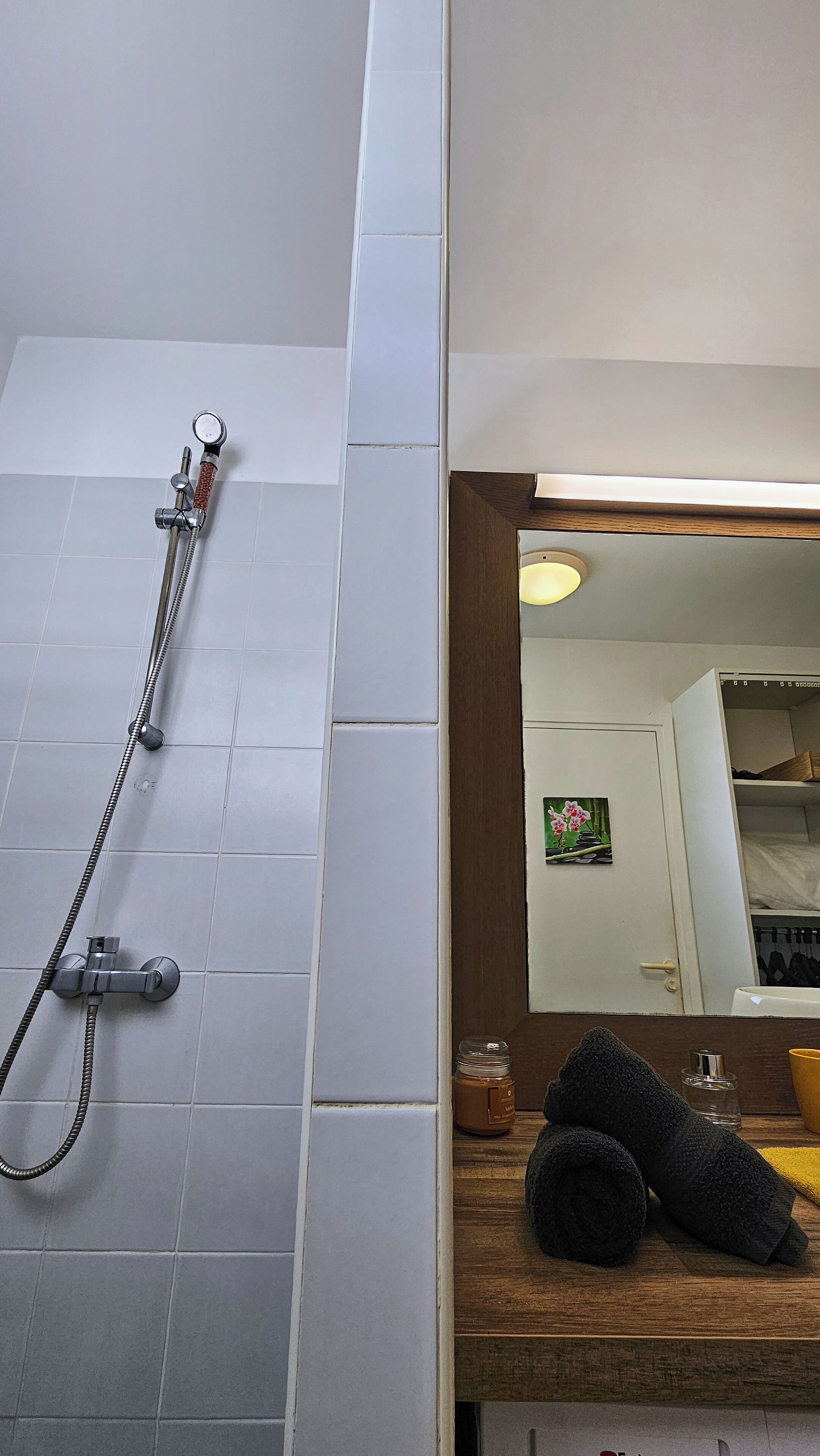 Um banheiro com chuveiro, uma prateleira de madeira com uma toalha e um espelho refletindo um quarto com um quadro.