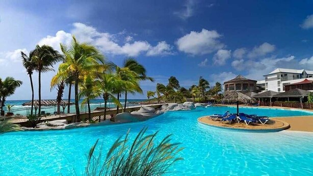 A resort pool with palm trees, a beach, and a building with a thatched roof.