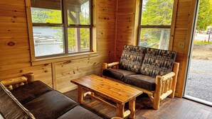 A wooden cabin interior with a sofa, a wooden table, and large windows offering views of a forest and a lake. - Hemlock - Big Moose Lakeside (Eagle Bay)