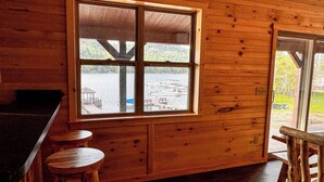 A wooden cabin interior with a large window overlooking a dock and boats. There are two wooden stools and a wooden chair. - Hemlock - Big Moose Lakeside (Eagle Bay)