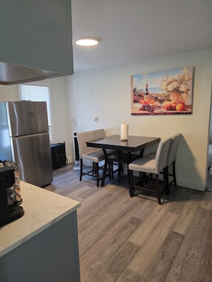 Una cocina con refrigerador, una mesa de comedor con sillas, un radiador y un cuadro en la pared.