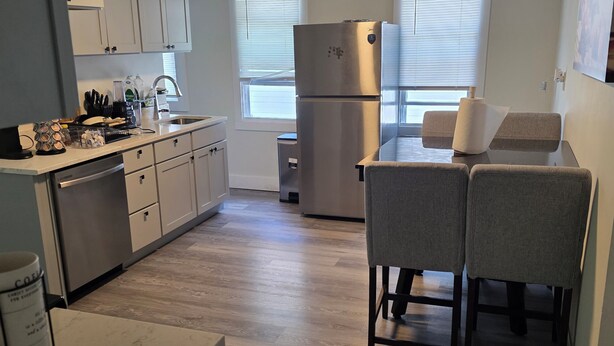A modern kitchen with white cabinets, a stainless steel refrigerator, a dining area with two chairs, and a coffee machine.